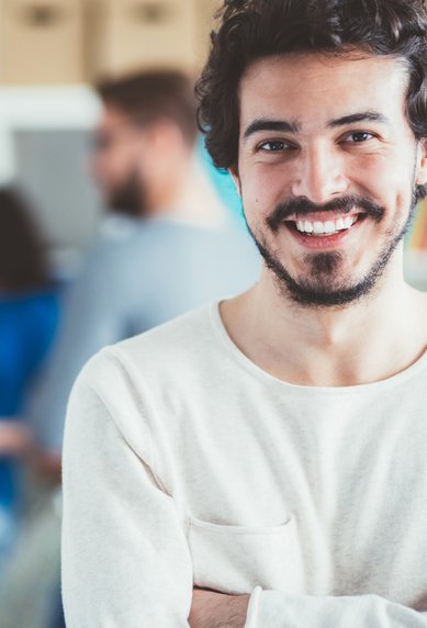 Ein junger Mann mit Bart lächelt und steht mit verschränkten Armen im Vordergrund, während im Hintergrund mehrere Personen an einer Wand arbeiten und sich unterhalten.